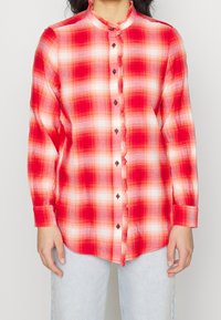 Kobieta w czerwono-białą kostkę, z długim rękawem, zapinana na guziki, z falbankami na brzegach, w połączeniu z jasnoniebieskimi dżinsami.