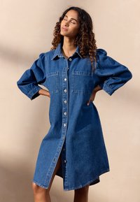 Denim-tyylinen paita mekko, puolipitkä, puffihuulisilla hihoilla, nappilista edessä ja kaksi rintataskua. Omistaa pehmeän, teksturoidun sinisen kankaan.