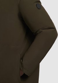 Olivengrøn pufferjakke med et struktureret stof, prydet med et fremtrædende rundt sort logo-patch på venstre ærme. Slim-fit design og sidelomme.