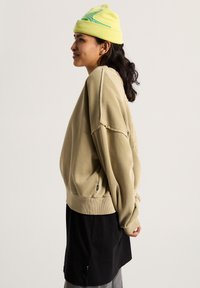 Felpa beige oversize con polsini e orlo a coste, caratterizzata da un dettaglio cucito. Abbinata a una gonna nera e un berretto giallo con accenti blu.