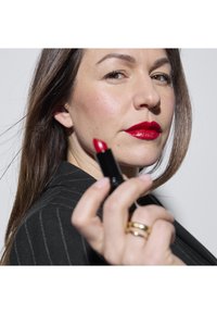 Musta huuliputki pidetään kädessä, ja siinä on kirkkaan punaista huulipunaa. Aiheella on pitkät ruskeat hiukset, mustapinnallinen bleiseri päällä ja punaiset huulet.