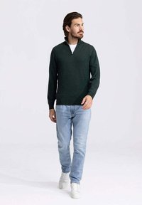 Donkergroene ritstrui in een gestructureerde brei, gecombineerd met lichtblauwe jeans en witte sneakers. Model staat tegen een lichte achtergrond.