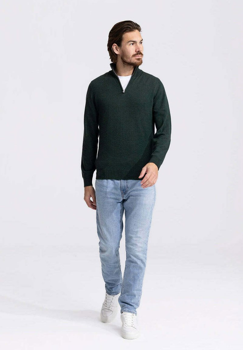 Donkergroene ritstrui in een gestructureerde brei, gecombineerd met lichtblauwe jeans en witte sneakers. Model staat tegen een lichte achtergrond.