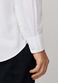 Camisa blanca de manga larga con collar clásico y puños abotonados, que presenta una textura sutil. Mano posicionada a la altura de la cintura, agarrando una tela oscura.