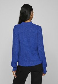 Blauwe, gebreide trui met een hoge hals en lange mouwen. De textuur is zacht en pluizig, met ribbelaccenten aan de onderrand en cuffs.