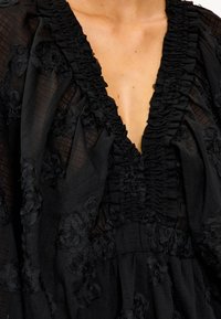 Blusa negra con un escote en V profundo, cuello con volantes y aplicaciones florales texturizadas. La tela es ligera y ligeramente transparente.