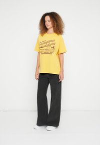 Yellow graphic t-shirt with "Sunseekers Paradise" print, paired with black wide-leg pants and white shoes. Smooth fabric and relaxed fit.