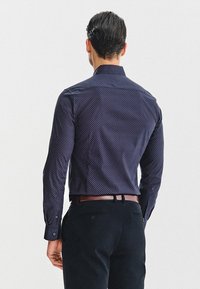 Mężczyzna z gładko zaczesanymi ciemnymi włosami, ubrany w wzorzystą ciemnoniebieską koszulę z guzikami, wsuniętą w czarne spodnie z brązowym paskiem, odwrócony plecami.