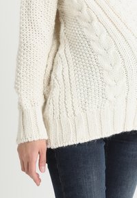 Sweterek w kolorze kremowym, dziany, z mieszanką wzorów ribbed i warkoczowych, o luźnym kroju i szerokich mankietach. Tekstura wydaje się miękka i gruba.