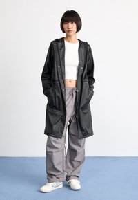 Svart, lång vattentät jacka med huva, med tryckknappar och sidofickor, i kombination med lösa grå cargobyxor och vita sneakers.