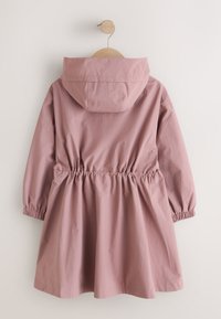 Imperméable à capuche rose poudré avec taille et poignets élastiques, suspendu sur un cintre en bois rond sur un fond blanc uni.