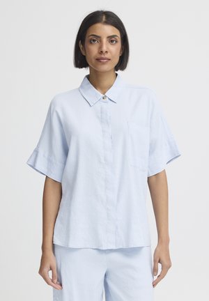 Woman with short dark hair wearing a light blue short-sleeve button-up shirt and matching light blue pants against a plain background.