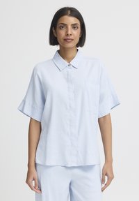 Femme aux cheveux courts et foncés portant une chemise à manches courtes bleu clair boutonnée et un pantalon bleu clair assorti, sur un fond uni.