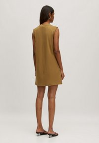 Ärmelloses, knielanges Kleid in einem Senfton, mit schlichtem Design, geradem Schnitt und Nahtdetail auf der Rückseite. Kombiniert mit schwarzen Sandalen.