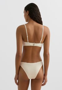 Krém színű bikini szett texturált anyagból, közép magas derekú alsó résszel és állítható pántokkal a felsőn, kapcsos záródással.