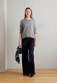 Mulher em pé no interior, vestindo um suéter cinza, calças largas escuras, segurando uma bolsa preta com um lenço listrado em preto e branco.