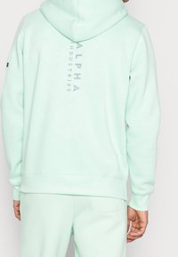 Felpa verde menta con cappuccio regolabile, logo grigio sulla schiena e polsini e orlo a coste. Tessuto morbido, design casual.