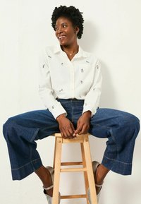 White button-up shirt with colorful embroidered designs, paired with wide-leg dark denim jeans and metallic silver heels, on a wooden stool.