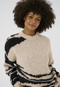 Pull en tricot beige avec des motifs en vagues noirs. Présente un col rond et des manches longues. Texture douce et coupe décontractée.