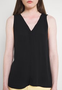 Žena v bezramenném černém topu s V-výstřihem a světlých kalhotách, zobrazená od ramen po boky na jednobarevném pozadí.