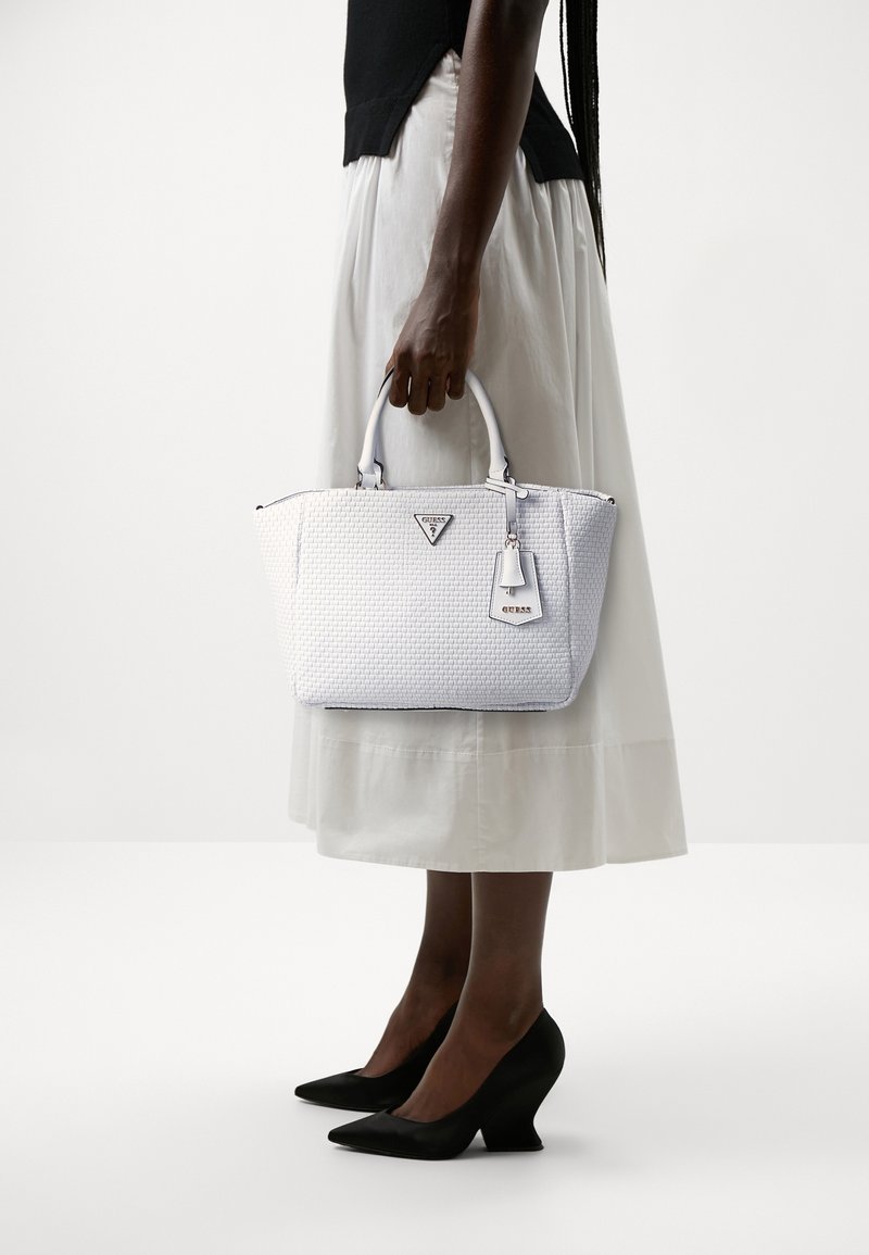 Personne portant une jupe blanche et un haut noir tient un sac à main tissé blanc avec double poignée et des chaussures à talons noires, debout sur un fond uni.