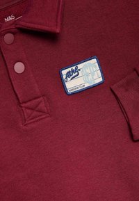 Burgundské polo tričko s dvoupoložkovým zapínáním, tkanou etiketou s modrým a bílým textem a texturovanou tkaninou. Akcentovaný límec a lem.