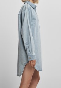 Valkblue denim-mekko, jossa on kauluskorsetti, pitkät hihat, nappi hihansuut ja kaareva helma. Tällä on yksi rintatasku.