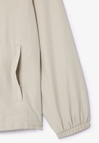 Beige, leichte Jacke mit weicher Textur, elastischen Bündchen und einer Seitentasche. Das Design zeichnet sich durch einen lockeren Schnitt und klare Linien aus.