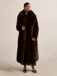 Lang brun pelsfrakk med bred krage, båret over en mørk kjole. Frakken har en myk, fluffy tekstur og en rett silhuett.