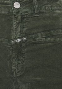 Pantalons en velours côtelé vert foncé avec une surface texturée, dotés d'une fermeture à bouton et d'une étiquette. Des poches visibles et des détails de couture sont présents.