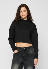 Svart crop-top genser med høy hals, avslappet passform og lange ermer. Kombinert med mørkegrå jeans med en falmet tekstur.