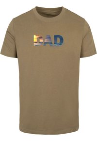 Beige bomulds t-shirt med ordet "DAD" i blå og solnedgangsbilleder, med teksten "i hele verden" i en mindre skrifttype.
