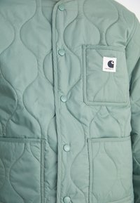 Gevoerde jas in lichtgroen met een textuurpatroon, voorzien van knopen aan de voorkant, een borstzak en een logo patch. Gemaakt van gladde stof.