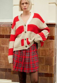 Femme blonde vêtue d'un cardigan oversized beige et rouge à rayures avec des boutons et d'une jupe écossaise rouge, se tenant devant un mur en carrelage marron.