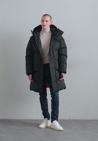 Manteau long noir rembourré avec un col haut, porté sur un pull à col roulé léger, associé à un jean foncé et des baskets blanches. Texture lisse, sans motifs.