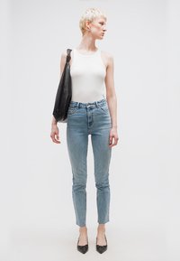 Άτομο που στέκεται με κοντά ξανθά μαλλιά, φοράει λευκή φανέλα χωρίς μανίκια, ψηλόμεσα μπλε τζιν, μαύρα μυτερά τακούνια και κρατά μια μεγάλη μαύρη τσάντα ώμου.