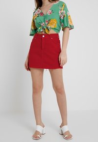 Top floreale verde con maniche corte, annodato in vita; gonna di denim rossa con orlo sfilacciato; sandali bianchi con tacco a blocco.