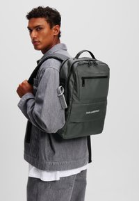 Rucsac verde închis cu două compartimente cu fermoar, bretele căptușite, accesorii gri și un logo pe față.