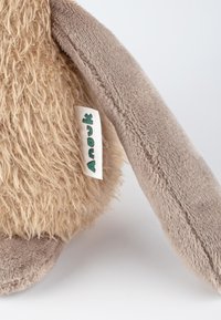 Plush toy with tan shaggy fur and soft beige limbs. Features a woven label with the text “Anouk” in green. Texture varies between materials.