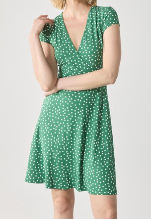 Femme portant une robe verte à manches courtes à pois blancs, taille cintrée et encolure en V, posant bras croisés et main près du cou.