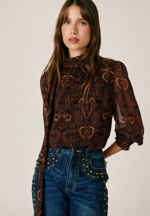 Mujer con cabello largo y castaño que lleva una blusa oscura con estampado y mangas abullonadas, y vaqueros azules de talle alto adornados con tachuelas doradas.