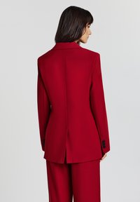 Roter taillierter Blazer mit strukturiertem Kragen, Einzelventil am Rücken und schwarzen Knöpfen an den Ärmeln. Glattes, leichtes Material.