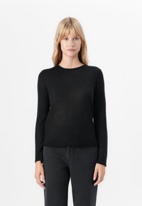 Woman with straight blonde hair wearing a black long-sleeve crew neck sweater and dark gray jeans against a white background.