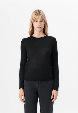 Woman with straight blonde hair wearing a black long-sleeve crew neck sweater and dark gray jeans against a white background.