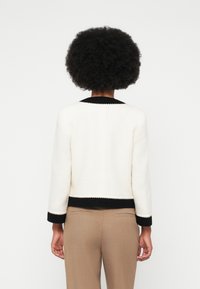 Veste blanche texturée avec des bordures noires au col et aux manches, portée sur un pantalon beige. Présente une silhouette carrée et des manches à la longueur du coude.
