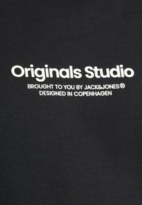 Fekete pamut anyag, amelyen merész fehér szöveg olvasható: "Originals Studio", "Brought to you by JACK&JONES®" és "Designed in COPENHAGEN."