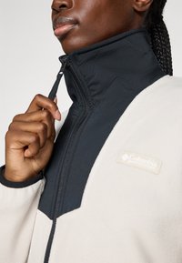 En person som drar opp glidelåsen på en to-farget Columbia fleecejakke med høy krage, og fletningen er synlig fra siden.