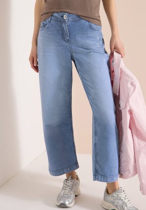 Kvinde iført blå- og hvidstribede bukser med brede ben og sølvfarvede sneakers, der holder en pink jakke i sin højre hånd.