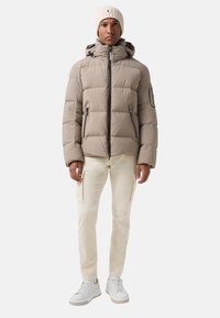Mann trägt eine beige Daunenjacke mit Kapuze, cremefarbene Cargo-Hosen, weiße Sneakers und eine beige Strickmütze mit kleinem Logo.