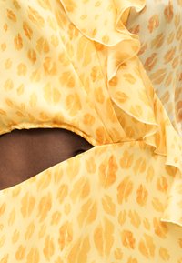 Κίτρινο ύφασμα με πορτοκαλί μοτίβο από αποτύπωμα χειλιών και λεπτομέρεια βολάν πάνω σε σκούρο δέρμα που αποκαλύπτει τμήμα με άνοιγμα-κλειδαρότρυπα.